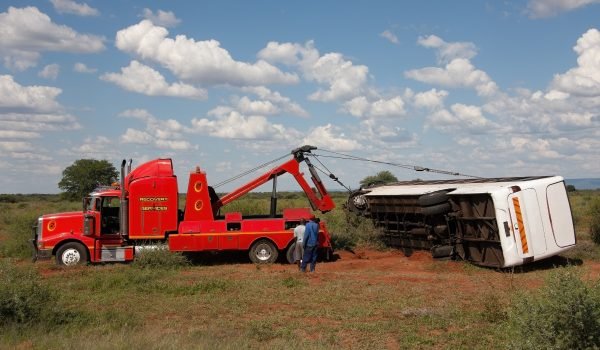 A winch out service is actively working to right an overturned vehicle in a rural landscape. Thick grasses and a bright blue sky provide the backdrop as two operators supervise the recovery process.